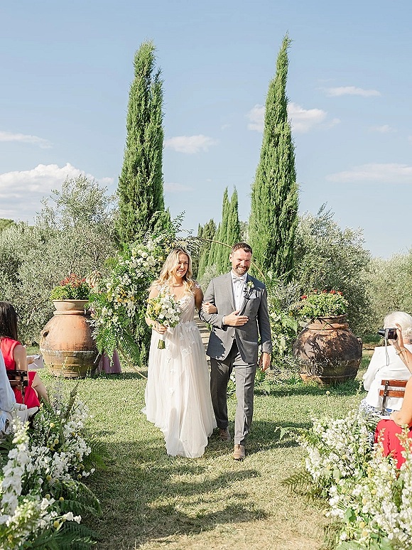 Ceremony recessional as bride and groom walk the aisle, bride holding a white bouquet beneath a floral arch in a sunny garden setting