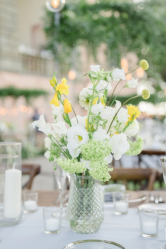 Wedding centerpiece with a glass vase of yellow and white flowers, candlelit table setting, and string lights in a garden reception background