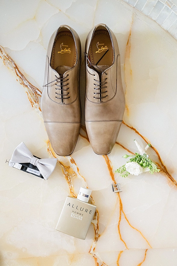 Groom details flatlay with tan dress shoes, white bow tie, boutonnieres, cologne bottle, and cufflinks on a marble surface