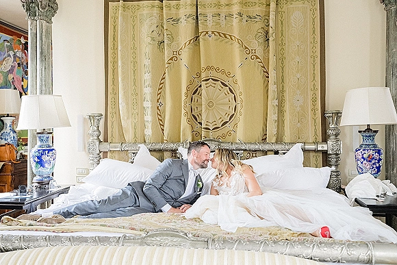 Wedding couple portrait of bride and groom on bed, kissing in a luxury bedroom with ornate canopy bed, lace train, and red heels