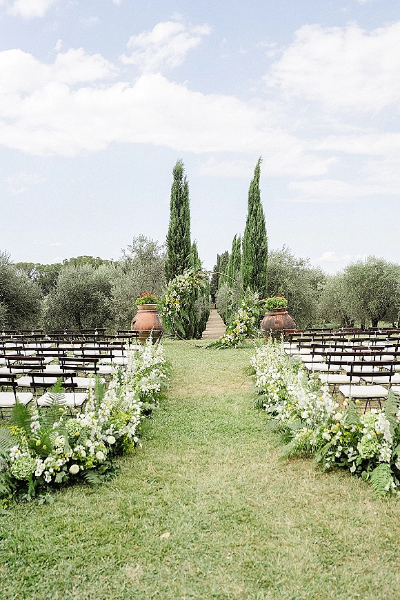 Ceremony setup for an outdoor wedding ceremony with a floral-lined aisle, wooden benches, and circular arch on a lawn by cypress trees