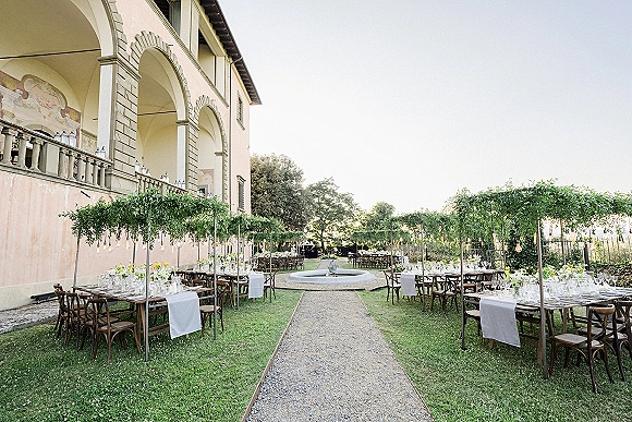 Outdoor reception setup with long banquet tables, white runners and floral centerpieces under greenery canopy and string lights in villa courtyard by fountain