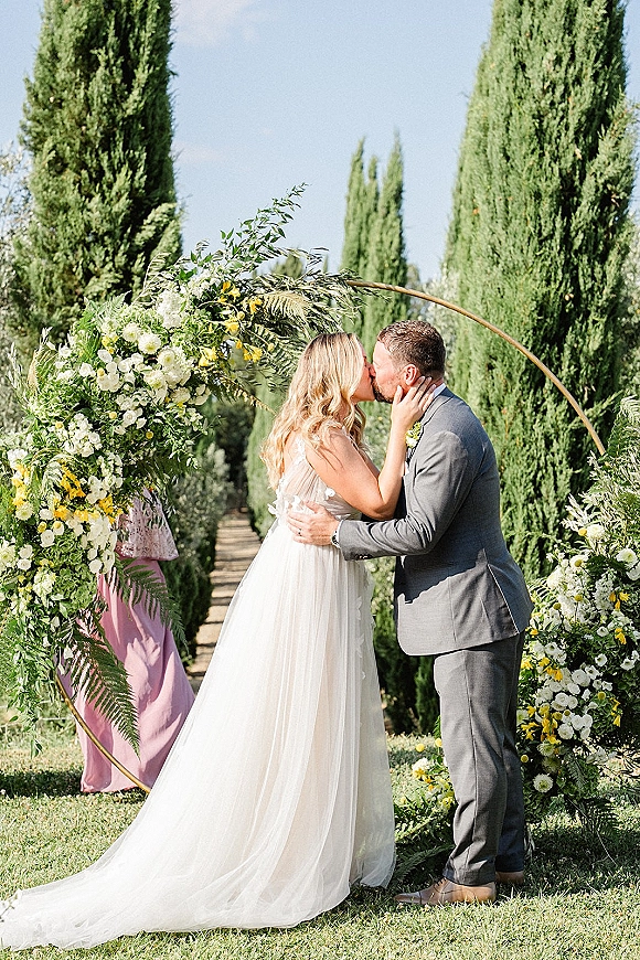 Wedding kiss portrait of bride and groom embracing under a floral circle arch with white and yellow flowers, cypress trees behind