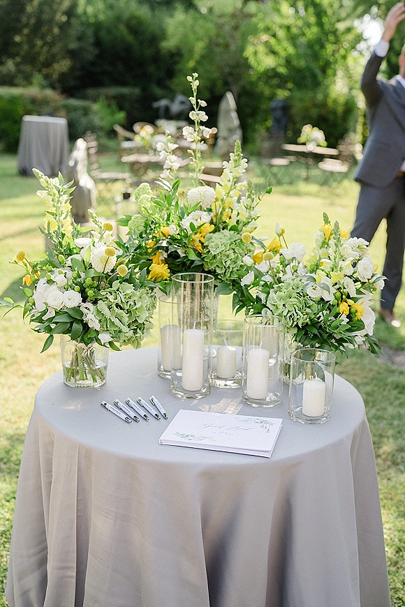 Wedding guest book table with floral arrangements and greenery, guest book and pens, and pillar candles in glass vases on a garden lawn