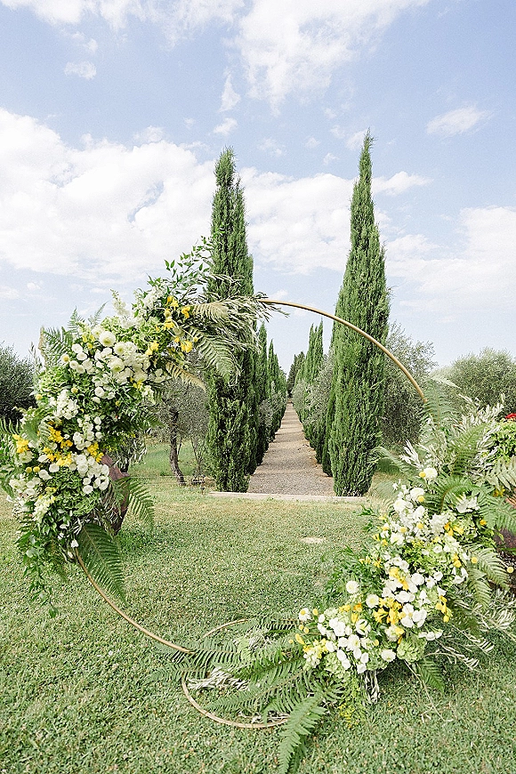 Wedding ceremony arch with a circular metal hoop and asymmetrical white and yellow flowers with greenery and ferns on a lawn by cypress trees