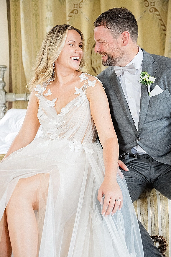 Couple portrait of bride and groom laughing on an upholstered bench, her lace bodice and his gray suit before neutral drapery background