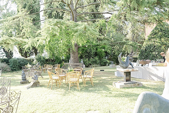 Outdoor cocktail area with rattan chairs and bistro tables, white linens and glassware beneath string lights on a garden lawn under a tree canopy