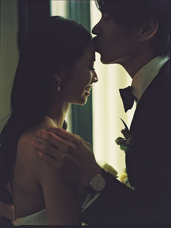 Wedding couple portrait of groom kissing bride’s forehead as she smiles, softly backlit by window light in an interior room