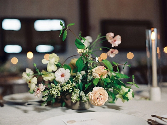 Reception centerpiece with wedding floral centerpiece of blush roses and greenery in a metal vase beside a taper candle on a candlelit table setting