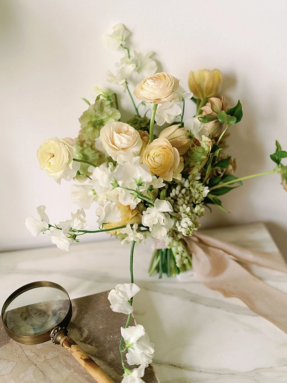 Bridal bouquet with white and yellow bouquet blooms of ranunculus and sweet peas, tied with blush ribbon on a marble surface near a magnifying glass