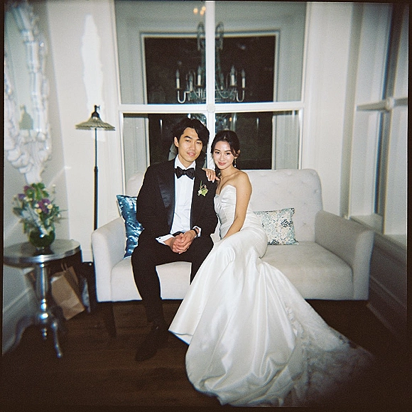 Couple portrait of bride and groom sitting on sofa, her strapless satin gown and his tuxedo, lit by window near chandelier in white room