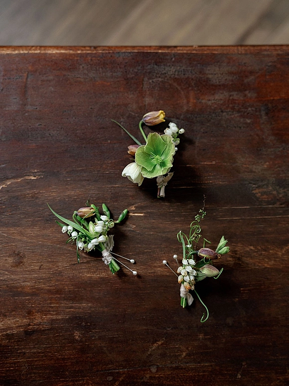 Wedding boutonnieres arranged in a flat lay with groom boutonniere, greenery, white floral tape, and pearl pins on a dark wood surface