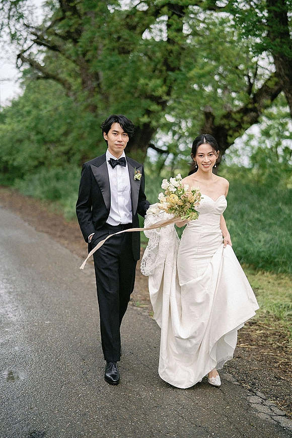 Couple portrait of bride and groom walking hand in hand on a tree-lined path, with bouquet ribbon and veil under an overcast sky