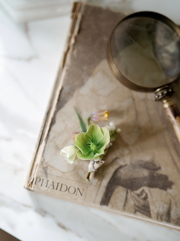 Boutonniere close-up of a green flower boutonniere with floral tape wrap resting on an antique book beside a magnifying glass on a white tabletop
