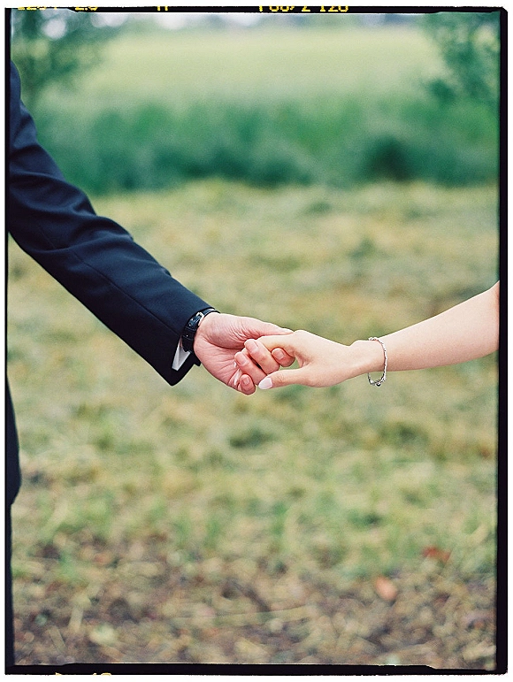 Couple holding hands in a wedding hand holding photo, interlocked fingers with groom’s suit jacket and wristwatch in a grassy field