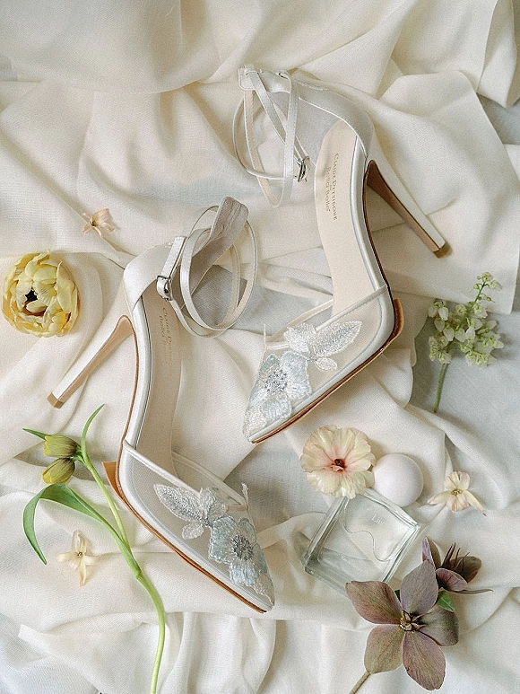 Bridal shoes in a wedding shoe flat lay, white strappy heels with ankle straps and floral appliqué beside perfume on draped fabric