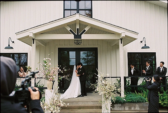Wedding kiss as bride and groom embrace on porch steps, bridal veil flowing, photographers nearby at white barn doors and florals