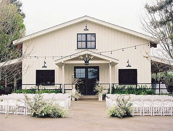 Ceremony setup with outdoor ceremony chairs in white folding chair rows, grounded aisle florals and string lights in front of a white barn