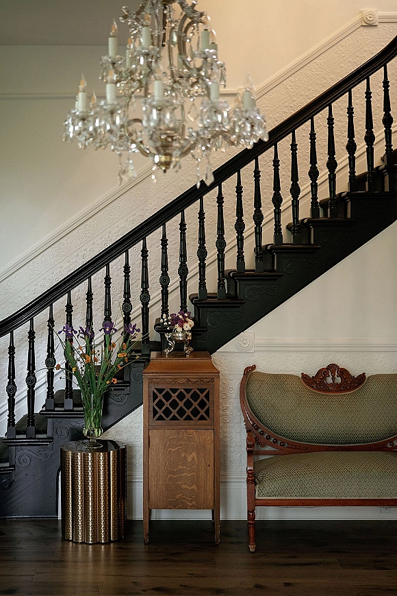 Wedding venue interior with a crystal chandelier above a grand staircase, floral pedestal arrangement, and vintage settee against textured white walls