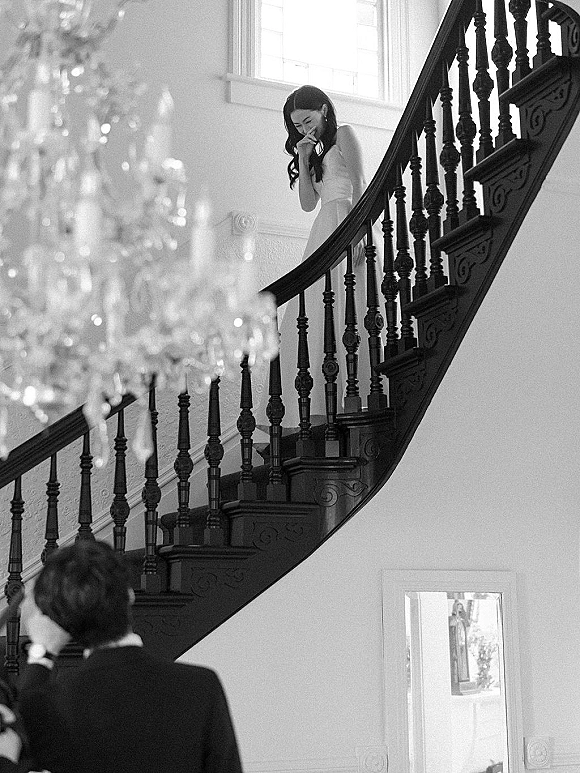 Wedding first look as bride comes down stairs in a bridal gown toward groom in suit jacket, under chandelier on bright interior staircase