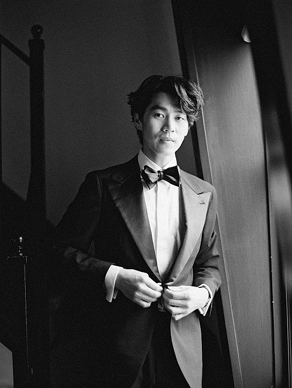 Groom portrait in a black tuxedo with satin lapels and bow tie, wearing a wristwatch, standing by a staircase railing in window light.