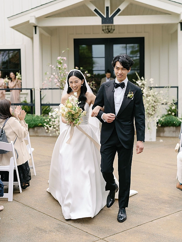 Wedding recessional as bride and groom walking past clapping guests, bride holding bouquet with long veil on an outdoor patio aisle