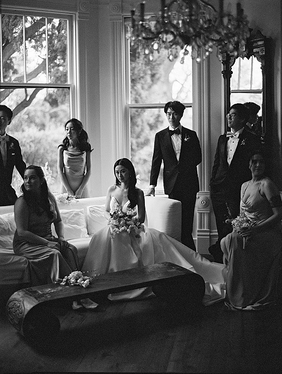 Wedding party portrait with bride on a sofa, bridesmaids holding bouquets and groom with groomsmen in tuxedos under a chandelier by large windows