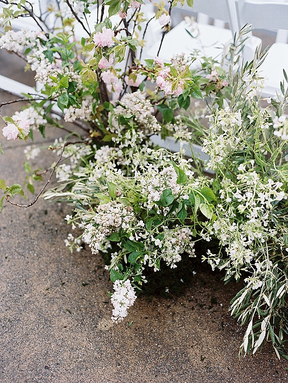 Wedding floral arrangement with greenery and pink-and-white flowering branches on a concrete floor beside white ceremony chairs