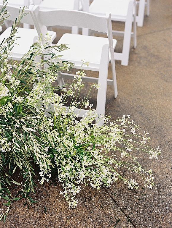 Ceremony aisle decor with white folding chairs lined along a concrete outdoor walkway, featuring low white flowers and greenery accents