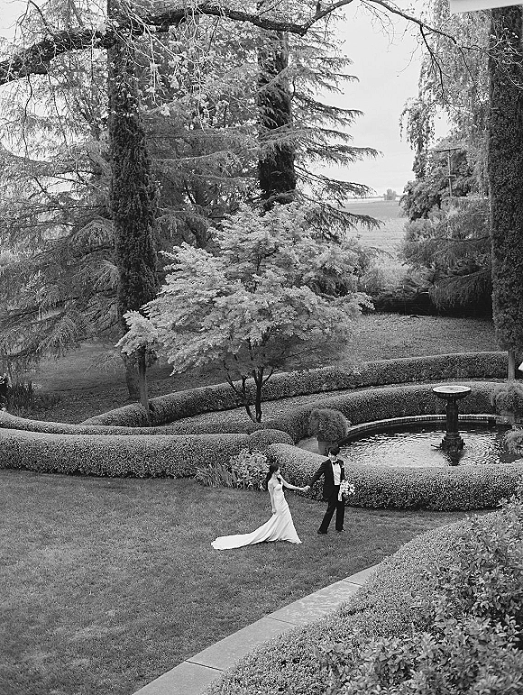 Couple portrait of bride and groom holding hands, her long train and white bouquet flowing beside his tuxedo in a formal garden fountain setting