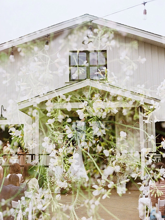 Outdoor ceremony setup with white chairs lining an aisle, greenery florals and string lights, set before a barn facade with guests seated