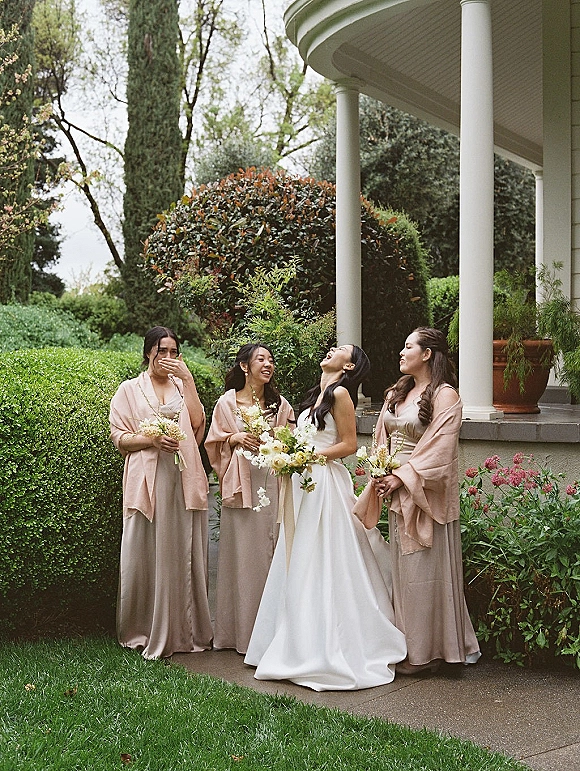 Bridesmaid group photo with bride with bridesmaids holding bouquets in neutral dresses and blush shawls on a garden lawn by white columns