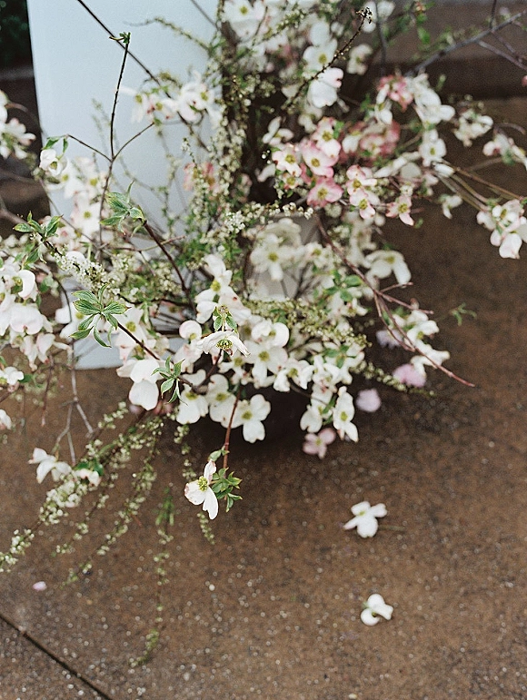 Wedding floral arrangement with blossom branches in a glass vase, mixing white and pink flowers with greenery on pavement with petals