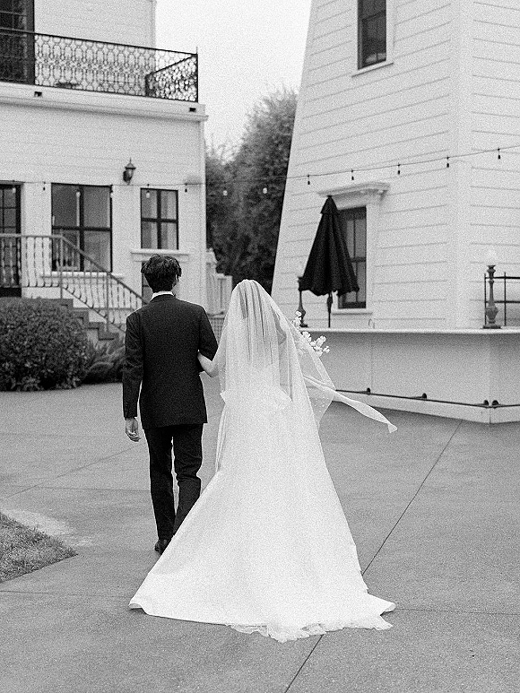 Wedding couple walking away, bride and groom from behind with veil blowing and long dress train under string lights by a white courtyard building
