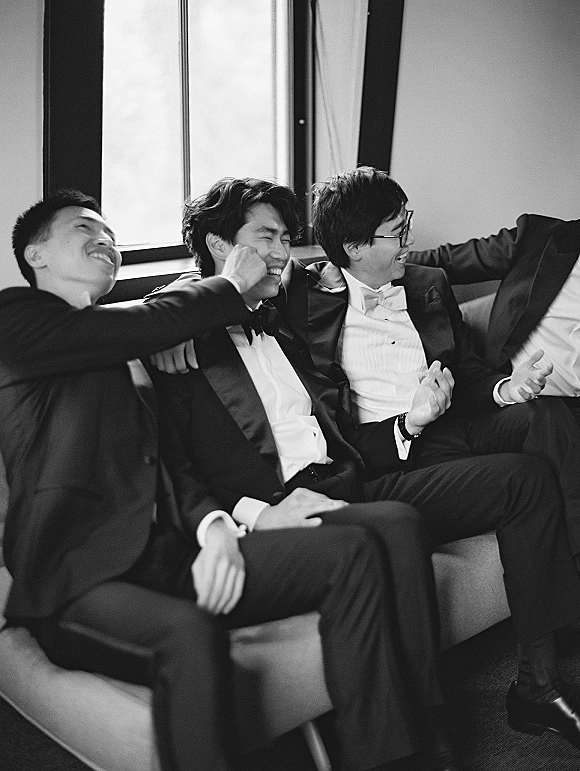 Groomsmen portrait of tuxedoed friends in bow ties laughing together on a couch by a bright window indoors, capturing candid camaraderie
