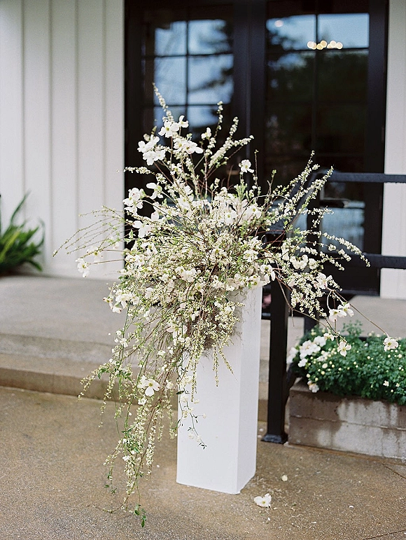 Wedding floral arrangement of white flowers and greenery with airy branches on a pedestal stand at a building entrance with glass doors