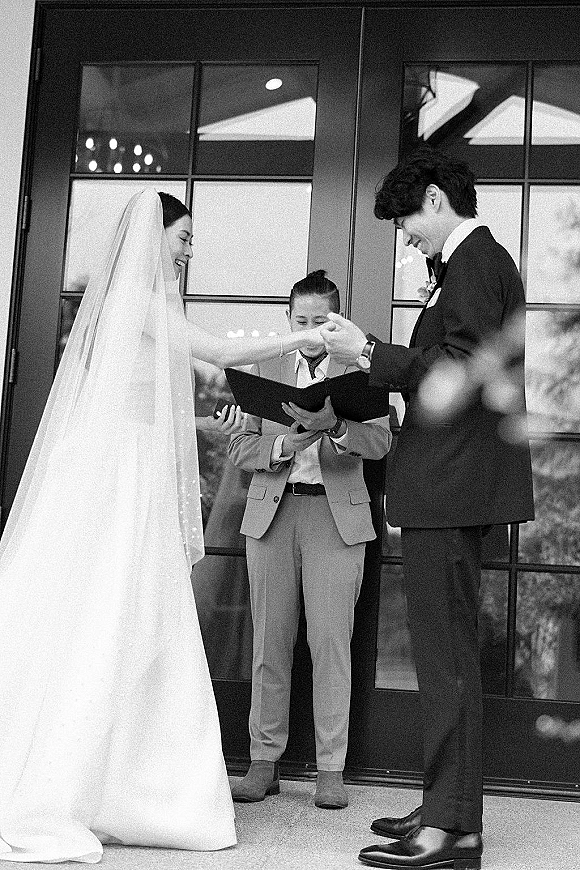 Wedding vows as bride and groom hold hands, her long veil trailing while officiant reads from a book before glass doors with reflections