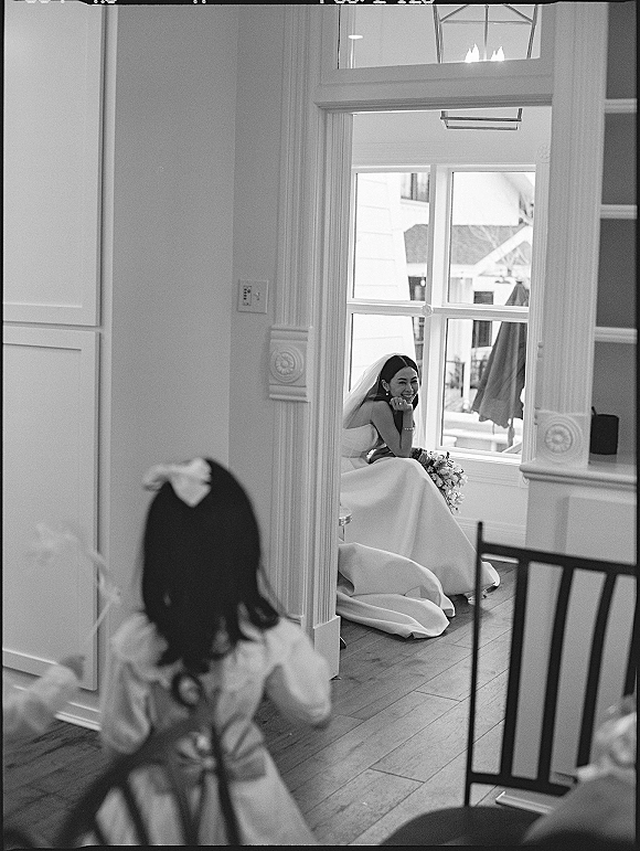 Bridal portrait of a bride sitting by window, laughing in a strapless dress with veil and bouquet, chandelier behind her