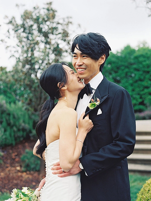 Wedding couple portrait of bride and groom hugging, sharing a forehead kiss beside garden greenery, bride in strapless dress with bouquet
