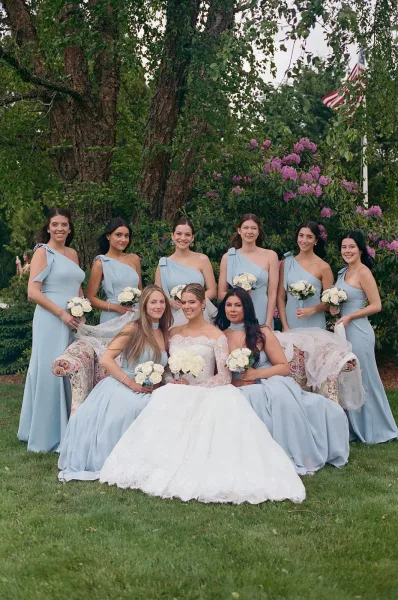 Bridal party portrait in dusty blue bridesmaid dresses posed on a garden lawn bordered by trees and flowering shrubs.