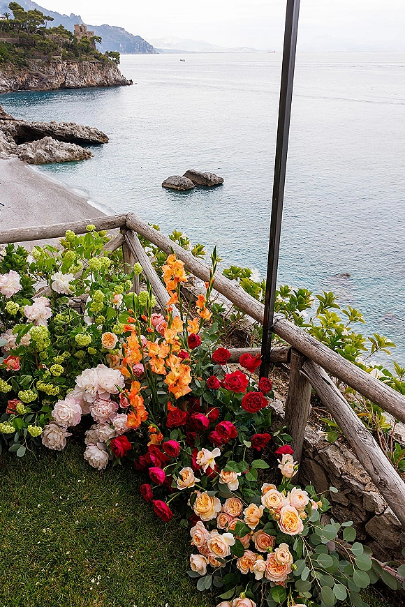 Wedding ceremony flowers in a lush ceremony floral installation with roses and peonies on a railing above a rocky ocean coastline and cliffs
