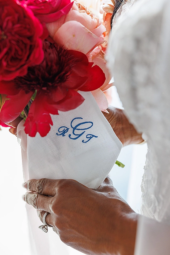 Wedding bouquet of red and blush flowers with a white wrap and blue monogram embroidery, held against soft bridal gown fabric