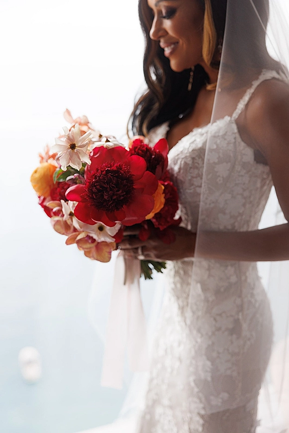 Bridal portrait of a bride holding bouquet in a lace wedding dress and veil, lit by a bright window with sky and water beyond
