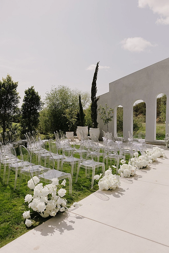 Ceremony setup with outdoor ceremony seating of clear acrylic chairs lining a white floral aisle runner before white stucco arches on a green lawn