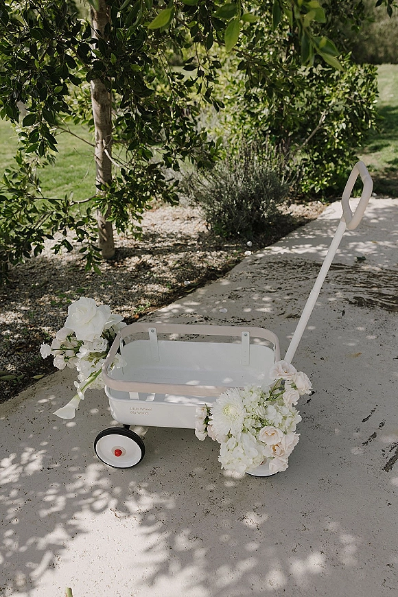 Flower girl wagon decorated with white flowers and greenery, ribbon and wheels, resting on a sunlit garden path with trees and shadows