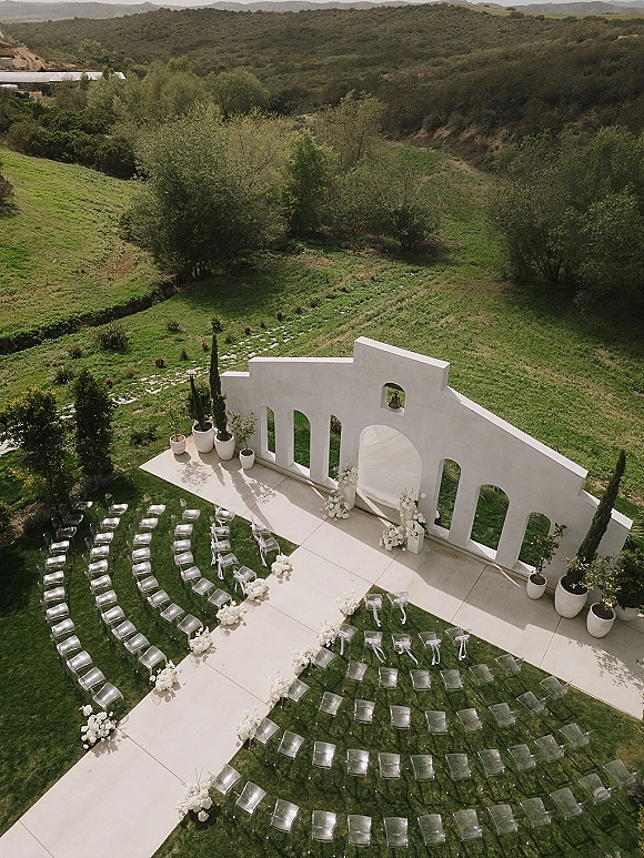 Ceremony setup with a white arched backdrop, clear acrylic chairs, and a white aisle runner lined with florals on a mountain-view lawn