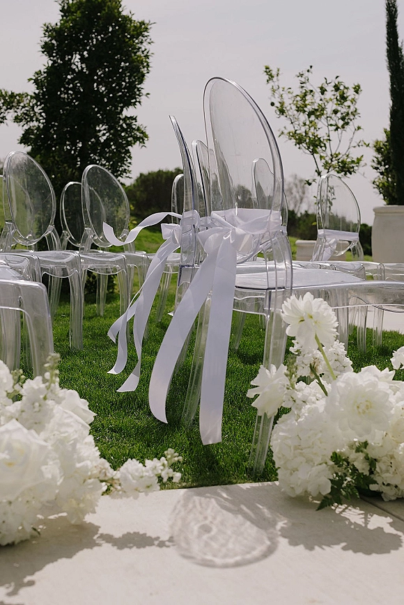 Ceremony aisle decor with clear acrylic ceremony chairs tied with white ribbons, flanked by white floral arrangements in glass vases on a lawn walkway