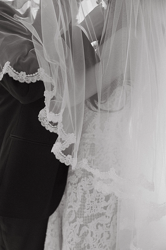 Wedding kiss as bride and groom kiss beneath a bridal veil, lace dress trim and groom’s dark suit in soft blurred background