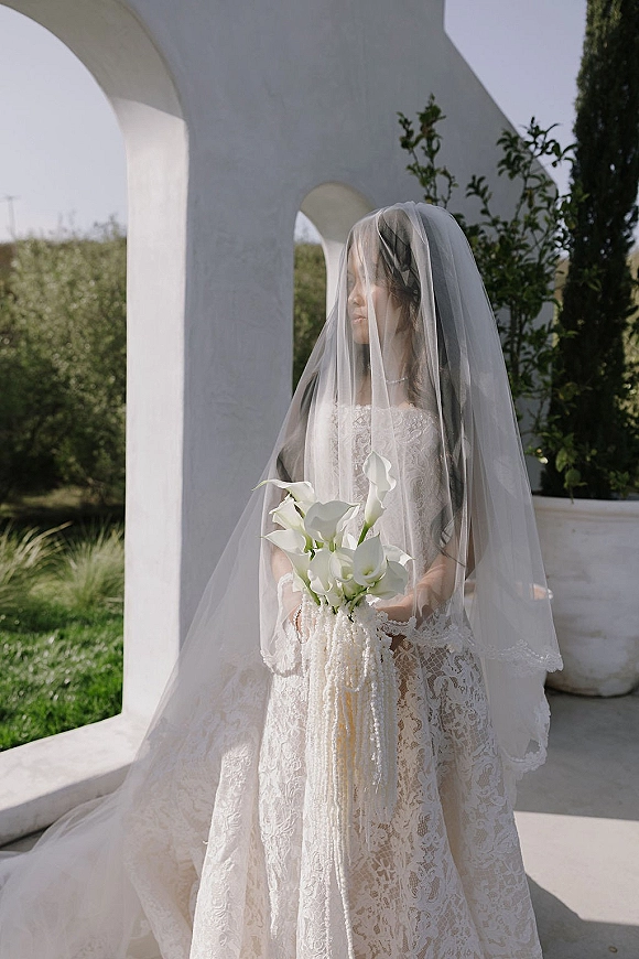 Bridal portrait of a bride with veil holding a calla lily bouquet, in a lace wedding dress under sunlit white stucco arches outdoors