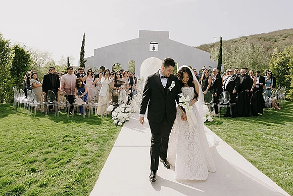 Wedding recessional as bride and groom walk the aisle, bride holding a white bouquet and veil flowing, guests seated before a white chapel facade